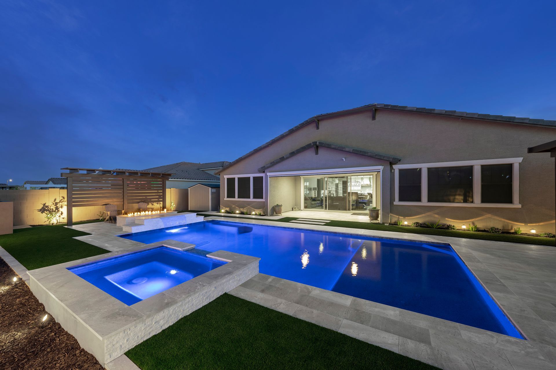 Backyard pool and spa at dusk with the house in the background. Blue water and sky, beige walls.