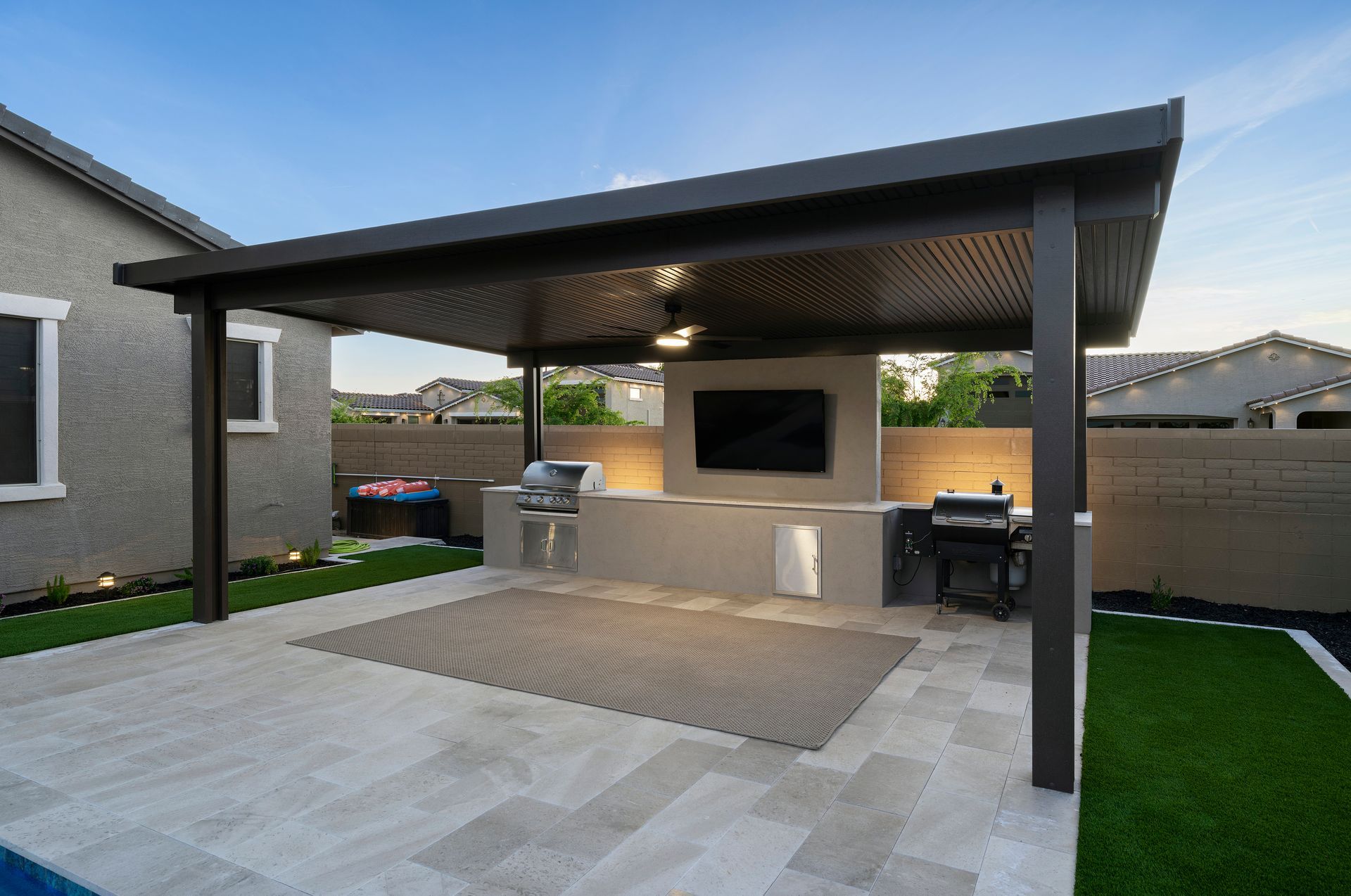 Outdoor kitchen patio with overhead dark-framed canopy, grill, TV, built-in counter, and rug.