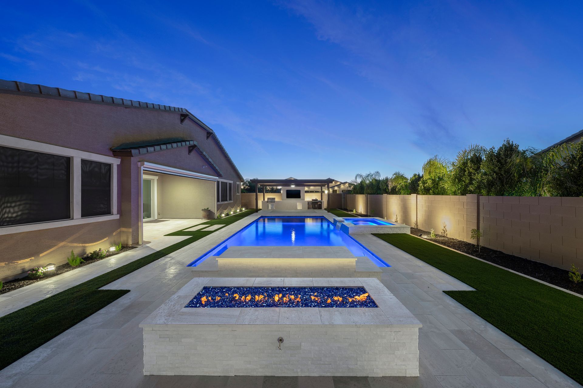 Backyard with a rectangular pool, fire pit, and a covered patio, illuminated by blue and warm lights at dusk.