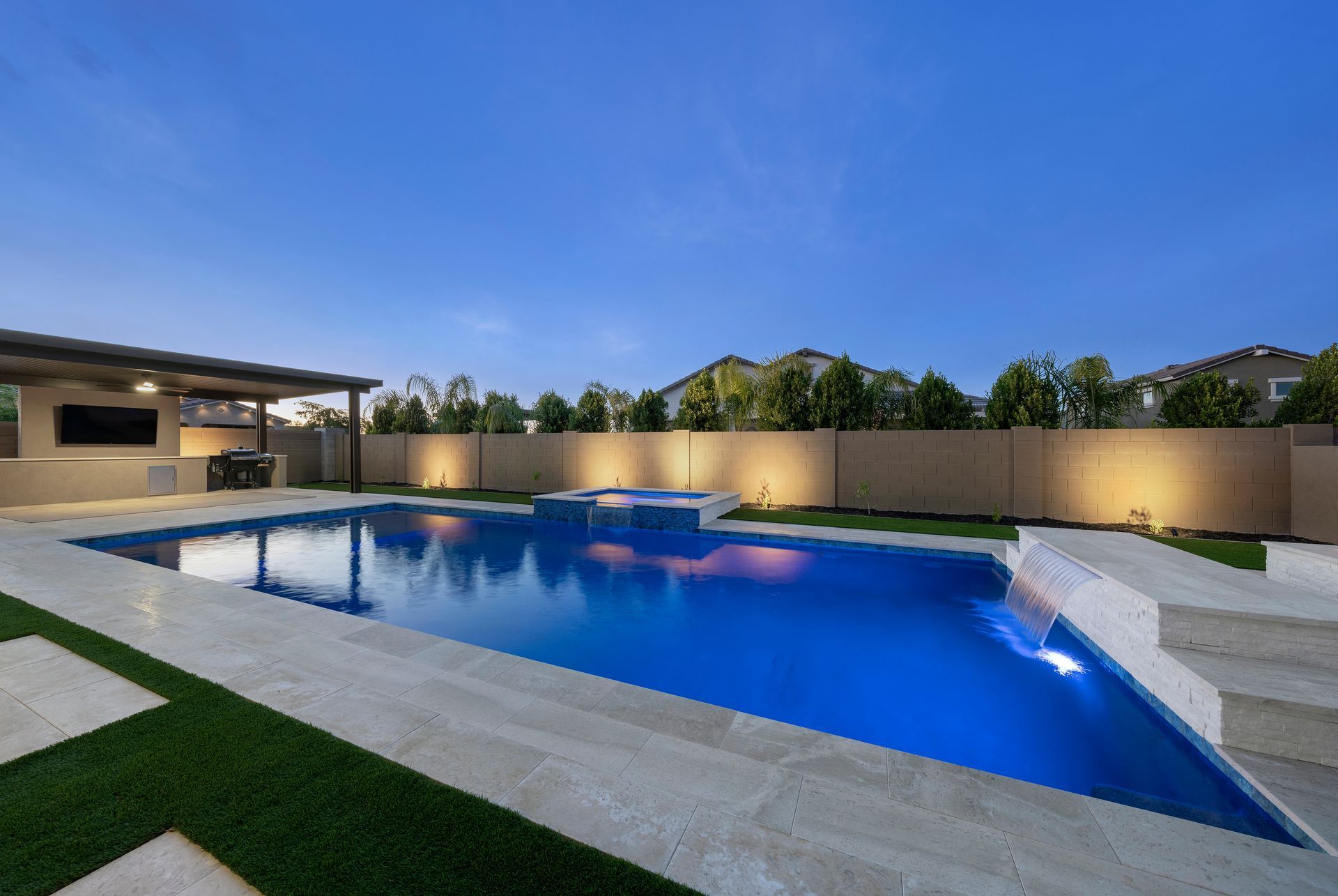Backyard pool illuminated at night. Blue water, stone patio, green turf, and outdoor kitchen.