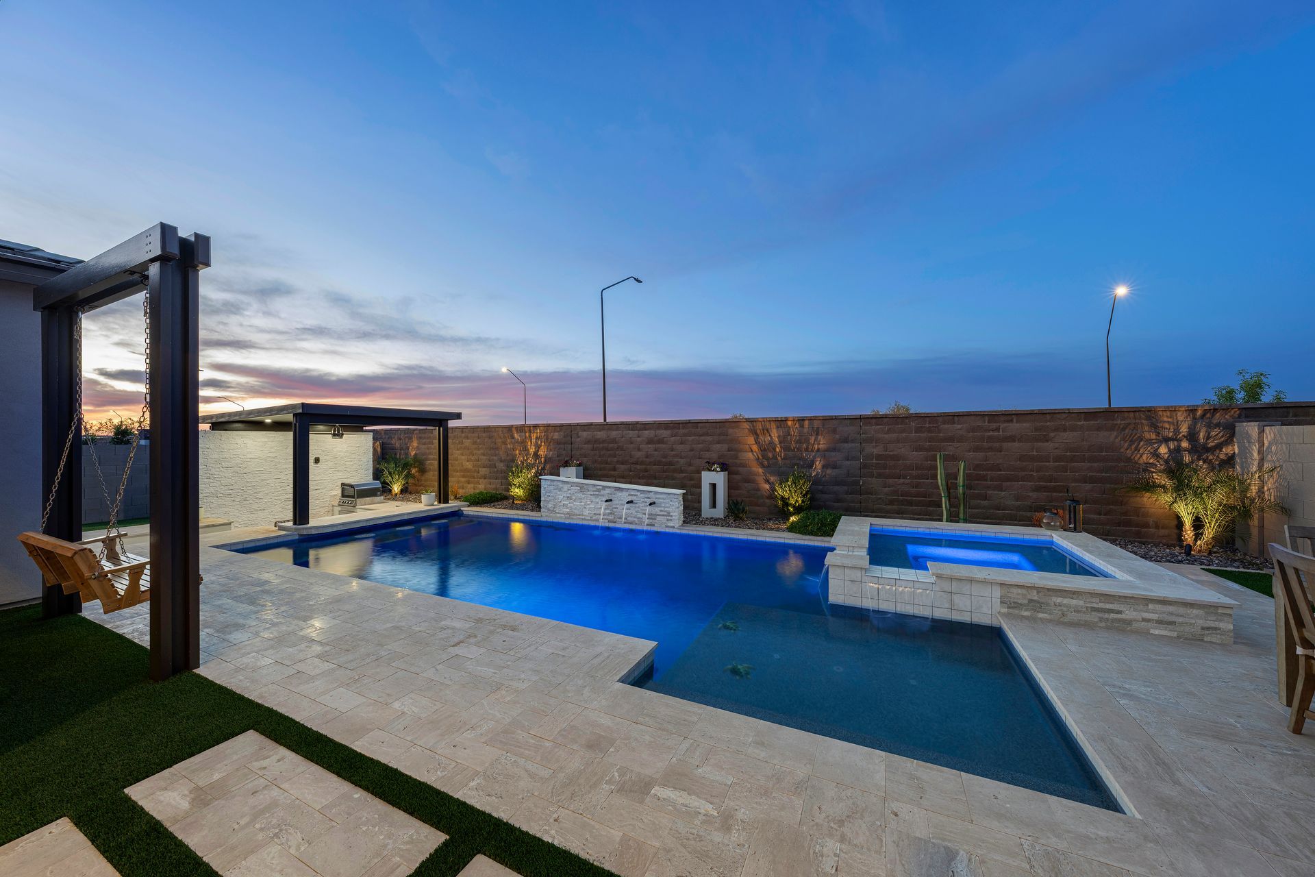 Pool and spa area with blue water, dusk sky, wooden pergola, and stone patio.