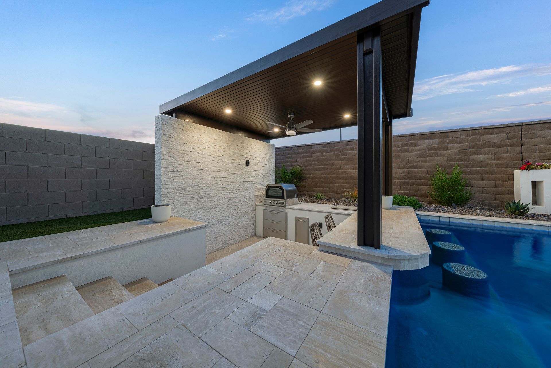 Poolside outdoor kitchen with bar seating under a dark brown pergola. Stone walls and a blue pool.