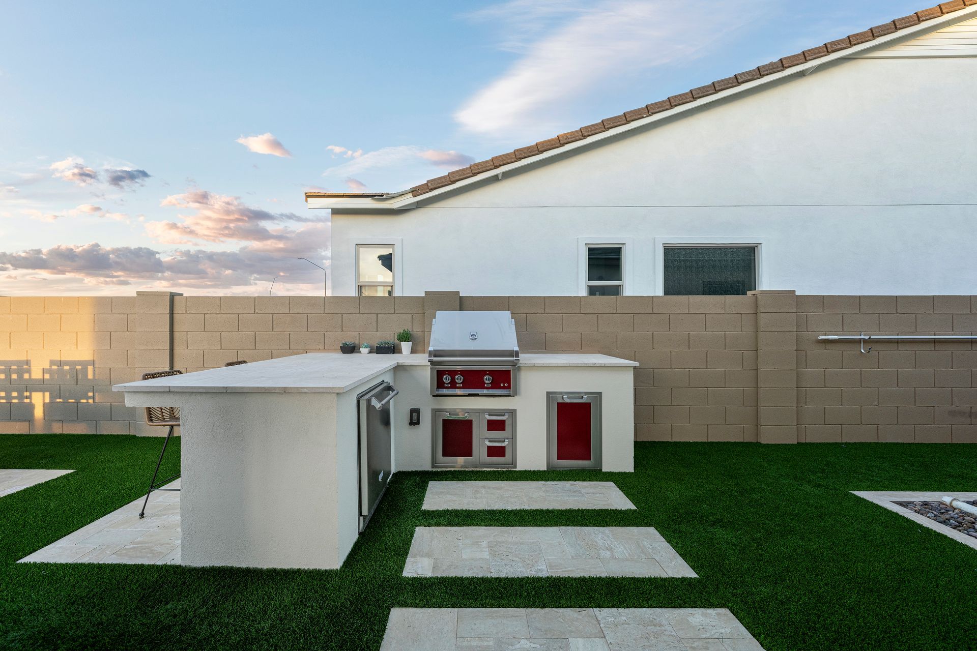 Outdoor kitchen with white countertops, grill, red doors, and a stone walkway on green turf, next to a white house.