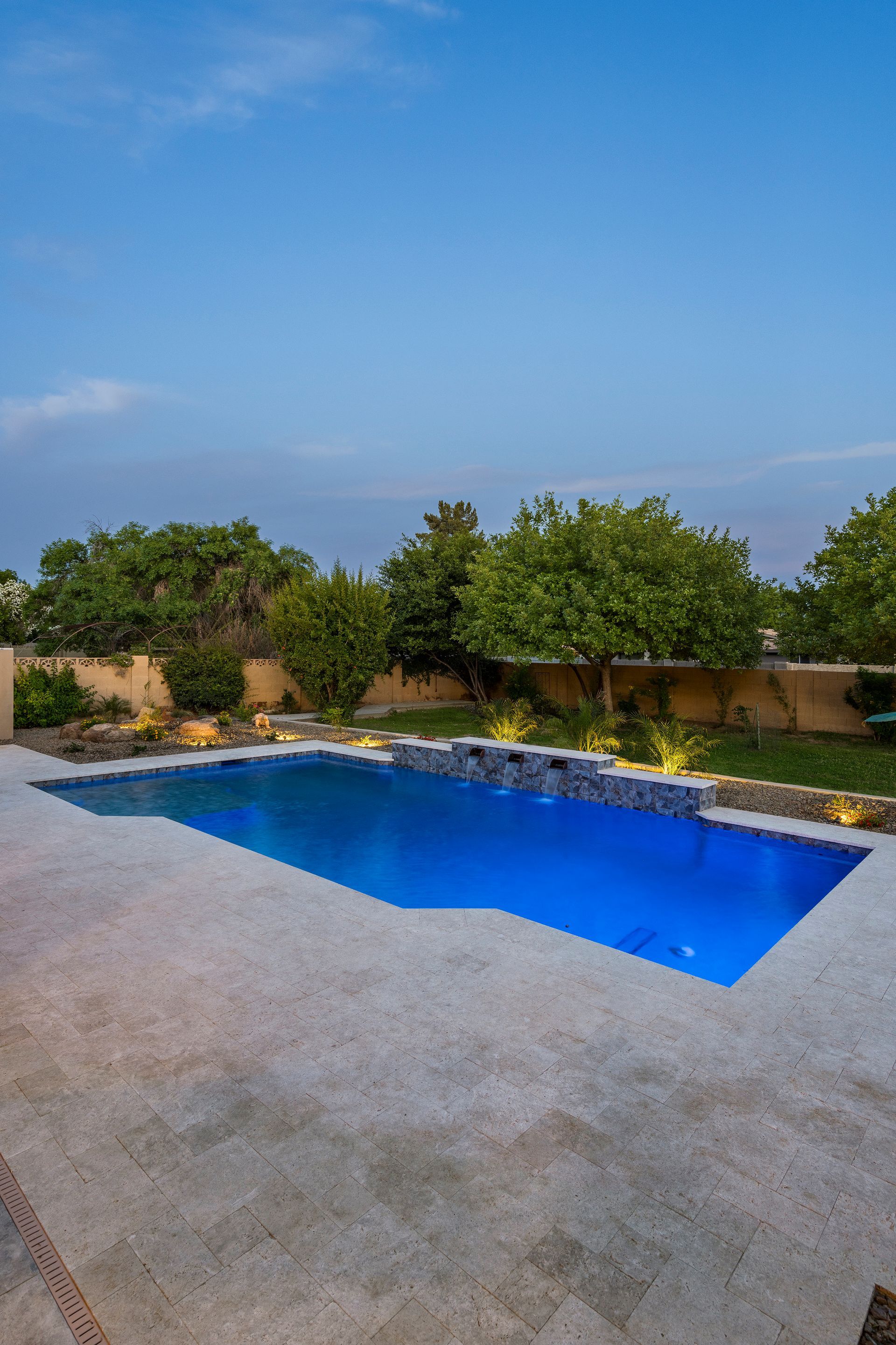 Swimming pool at dusk with blue lighting, surrounded by a stone patio and landscaping.