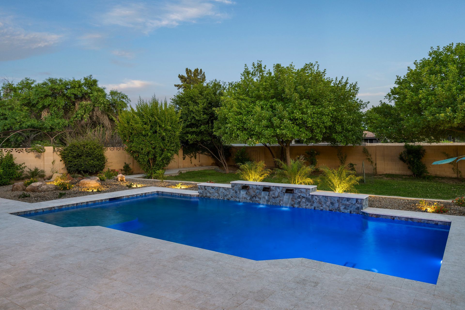 Pool in backyard, blue water, stone accents, surrounded by greenery and trees under a dusk sky.