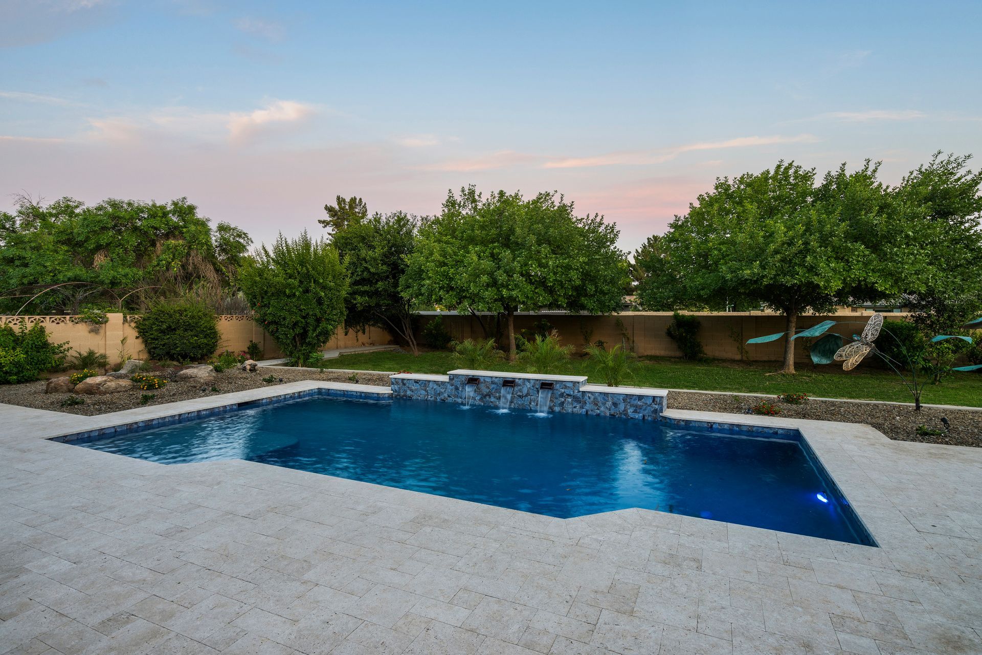 Backyard pool with stone deck, water feature, and trees under a colorful sky.