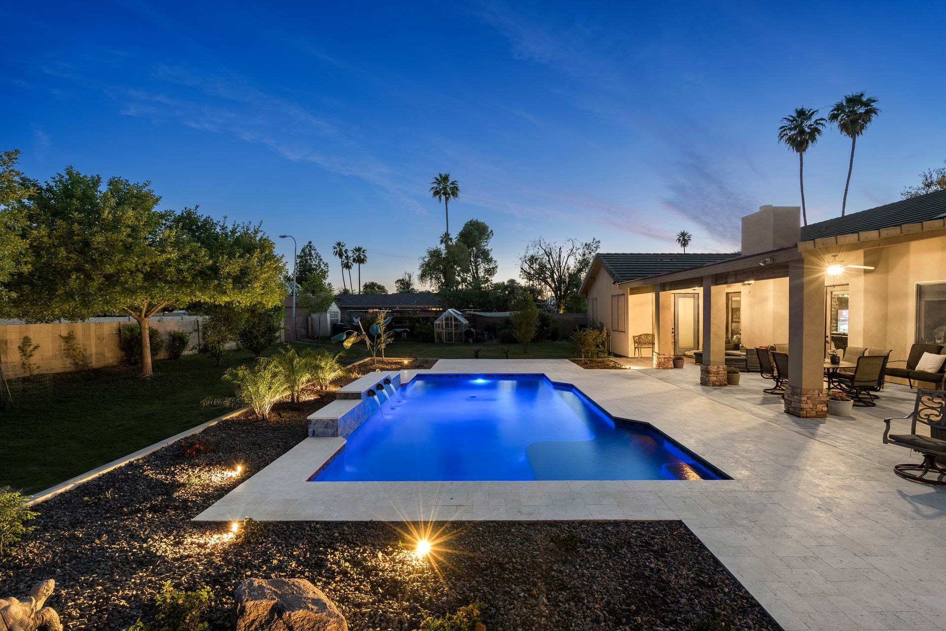Backyard with illuminated pool, patio, and house at dusk. Palm trees in background.