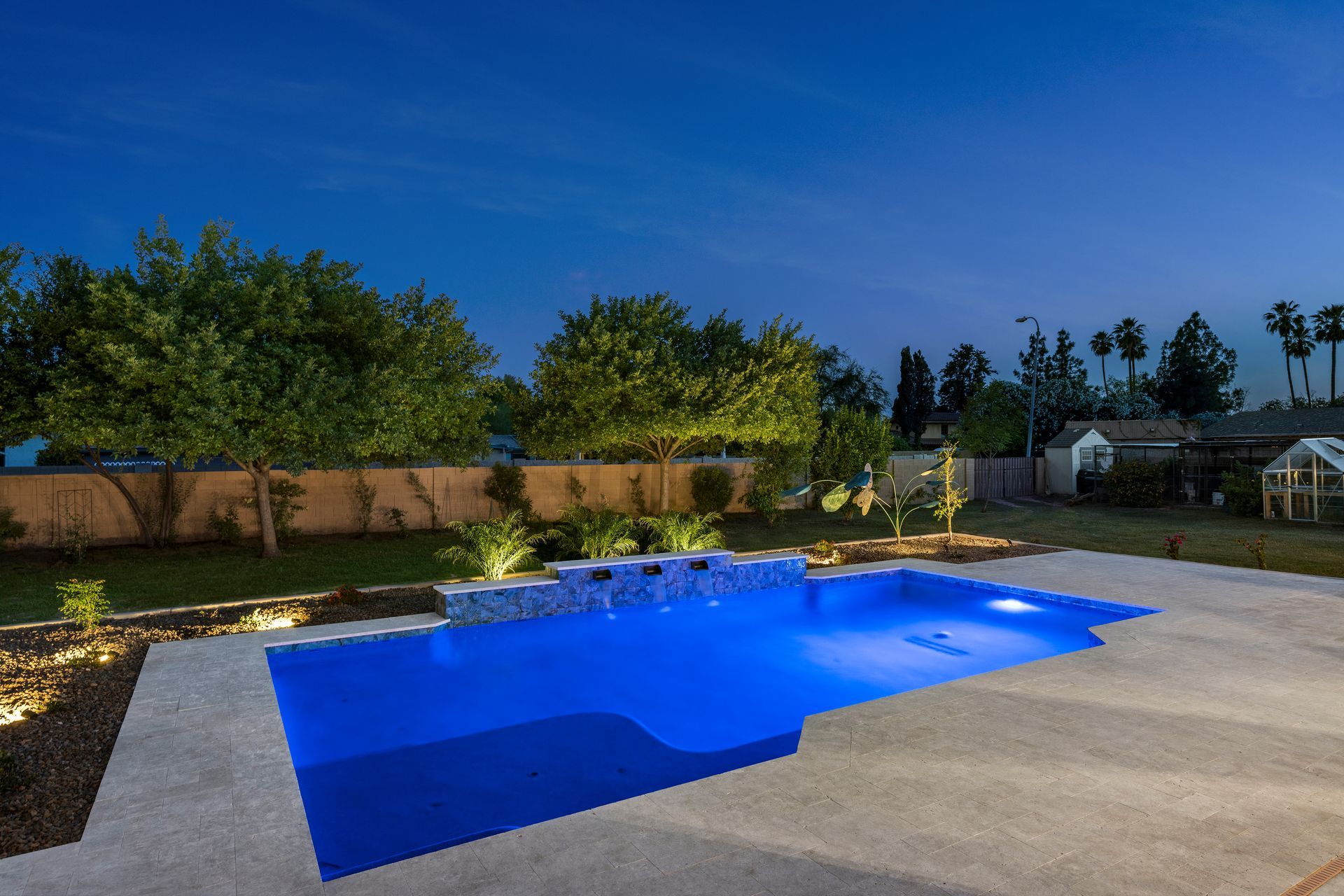 Blue-lit swimming pool at dusk, with landscaping, trees, and a clear sky in the background.
