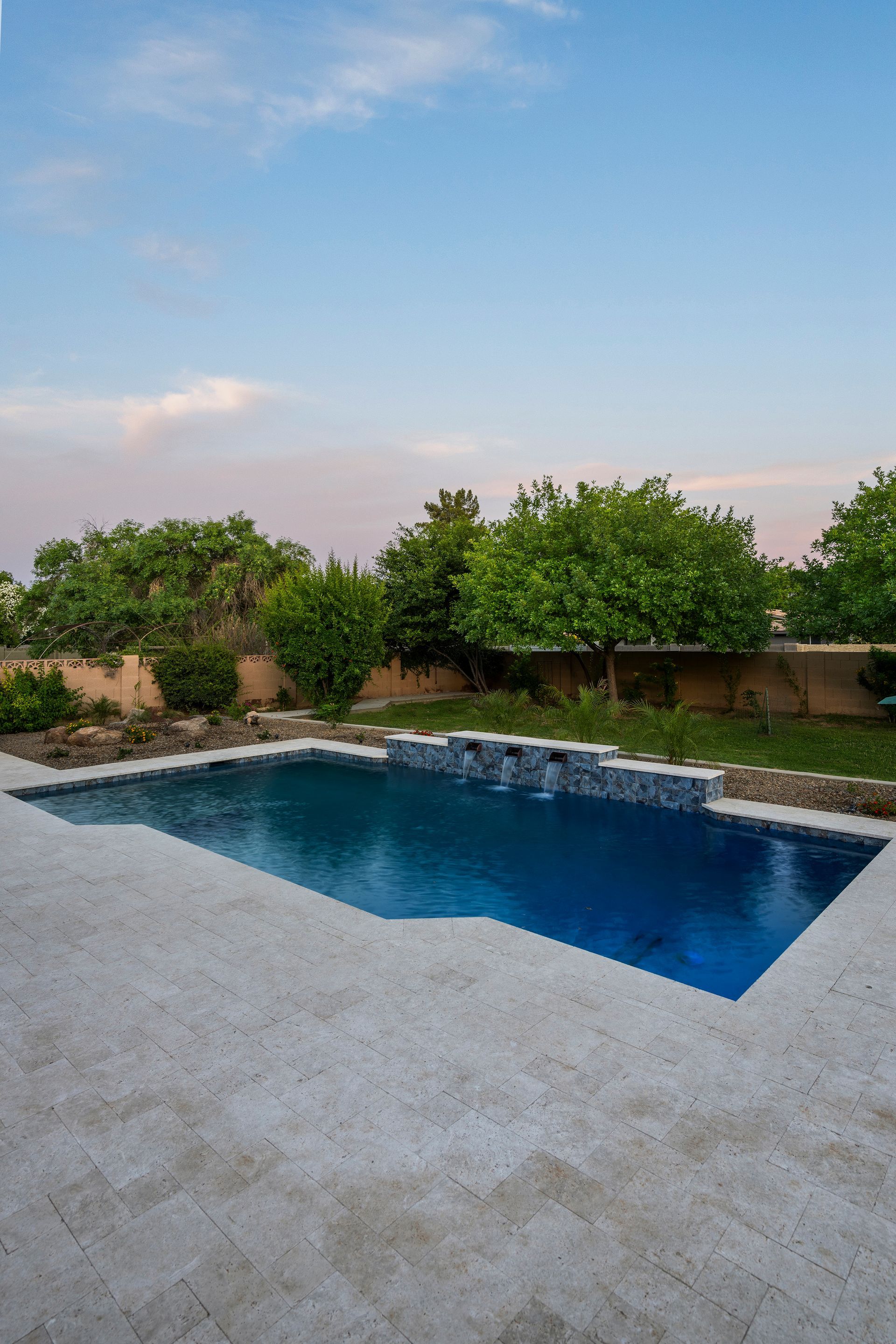 Pool with blue water surrounded by stone patio, lush greenery, and blue sky.
