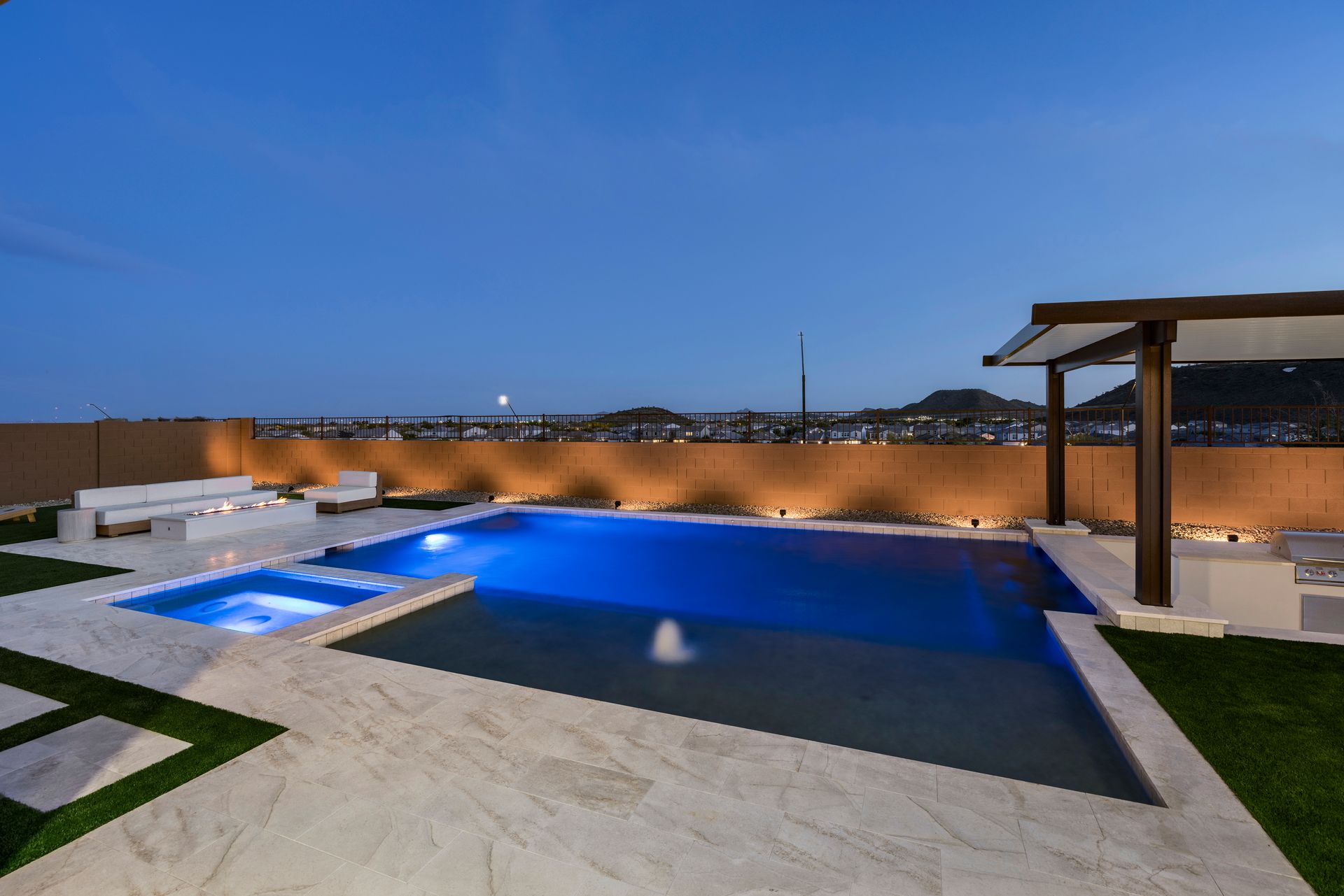 Night view of a backyard with a pool and jacuzzi, a pergola, and white seating.