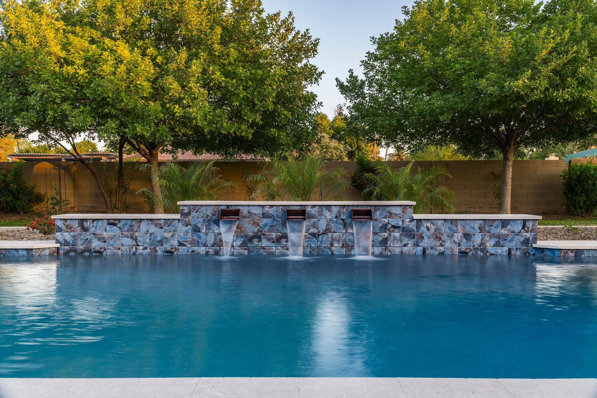 Swimming pool with stone waterfall feature, turquoise water, and trees in the background.
