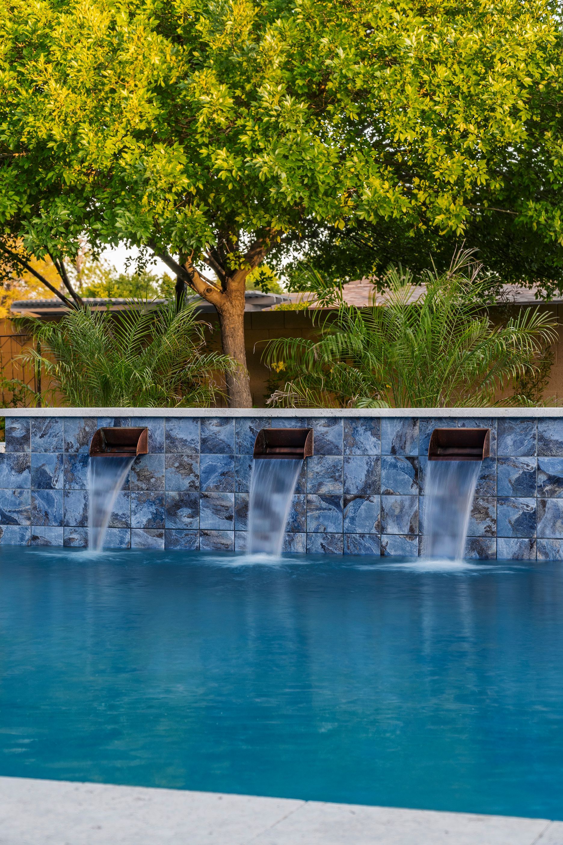 Pool with three water features against a stone wall, tree in background.