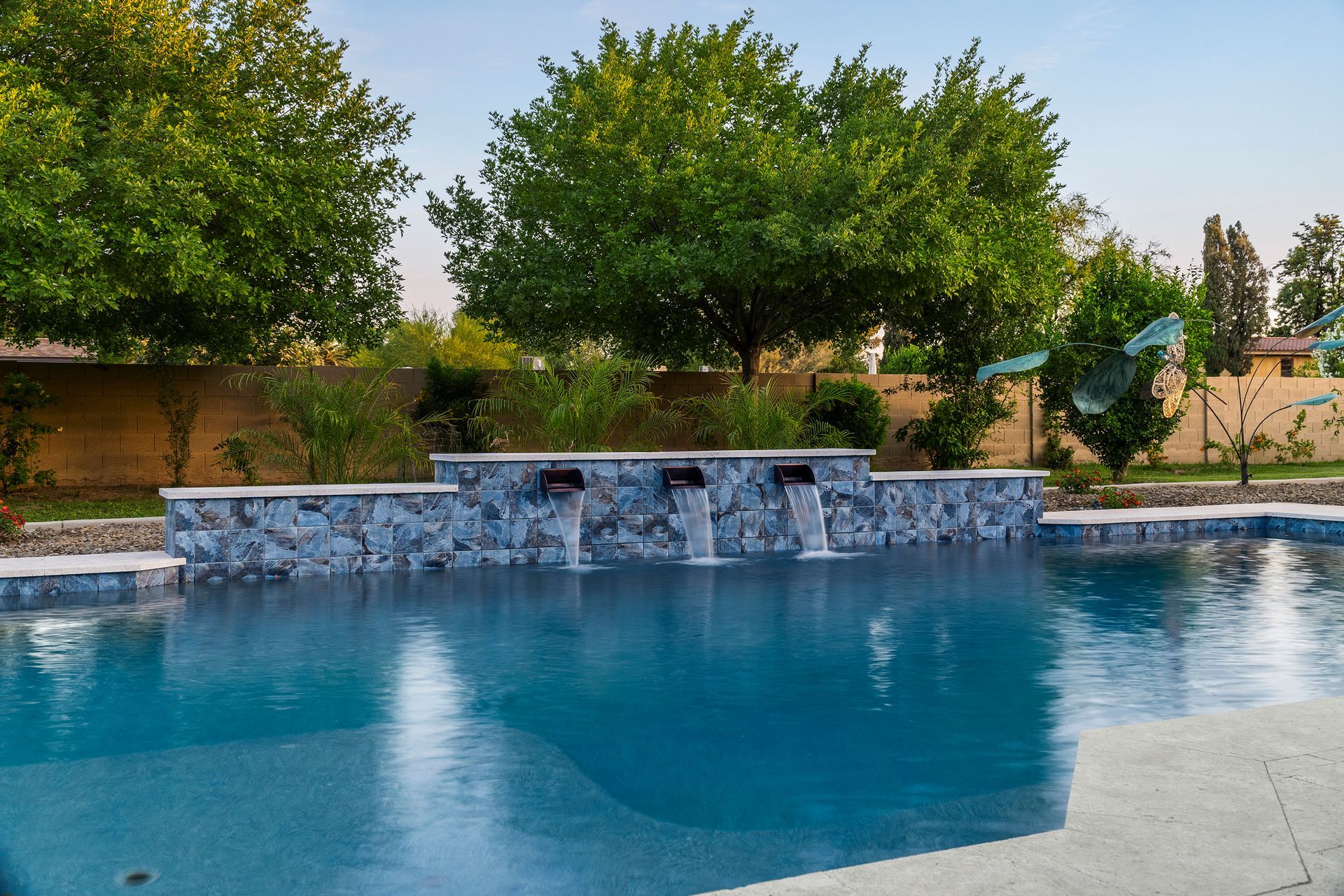 Swimming pool with a stone water feature, blue water, green trees, and tan wall backdrop.