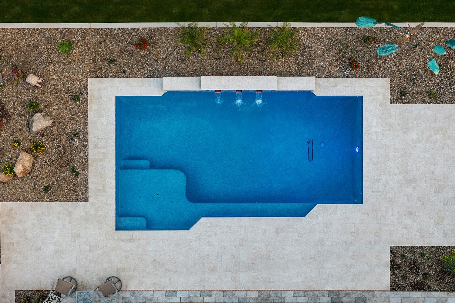 Aerial view of a rectangular blue swimming pool with steps, surrounded by light stone and landscaping.