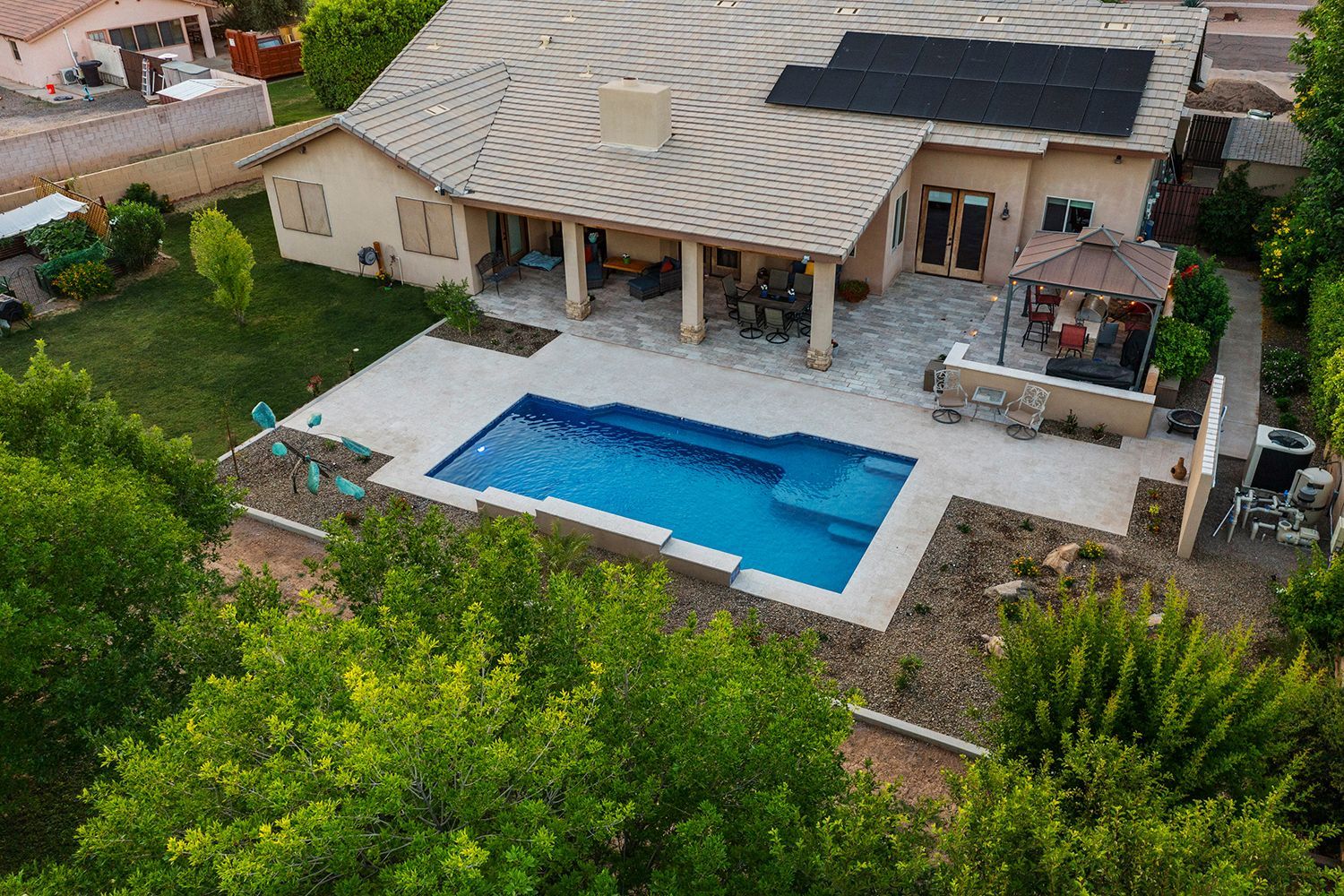 Aerial view of a home with a blue pool, patio, and solar panels. Green trees surround the property.