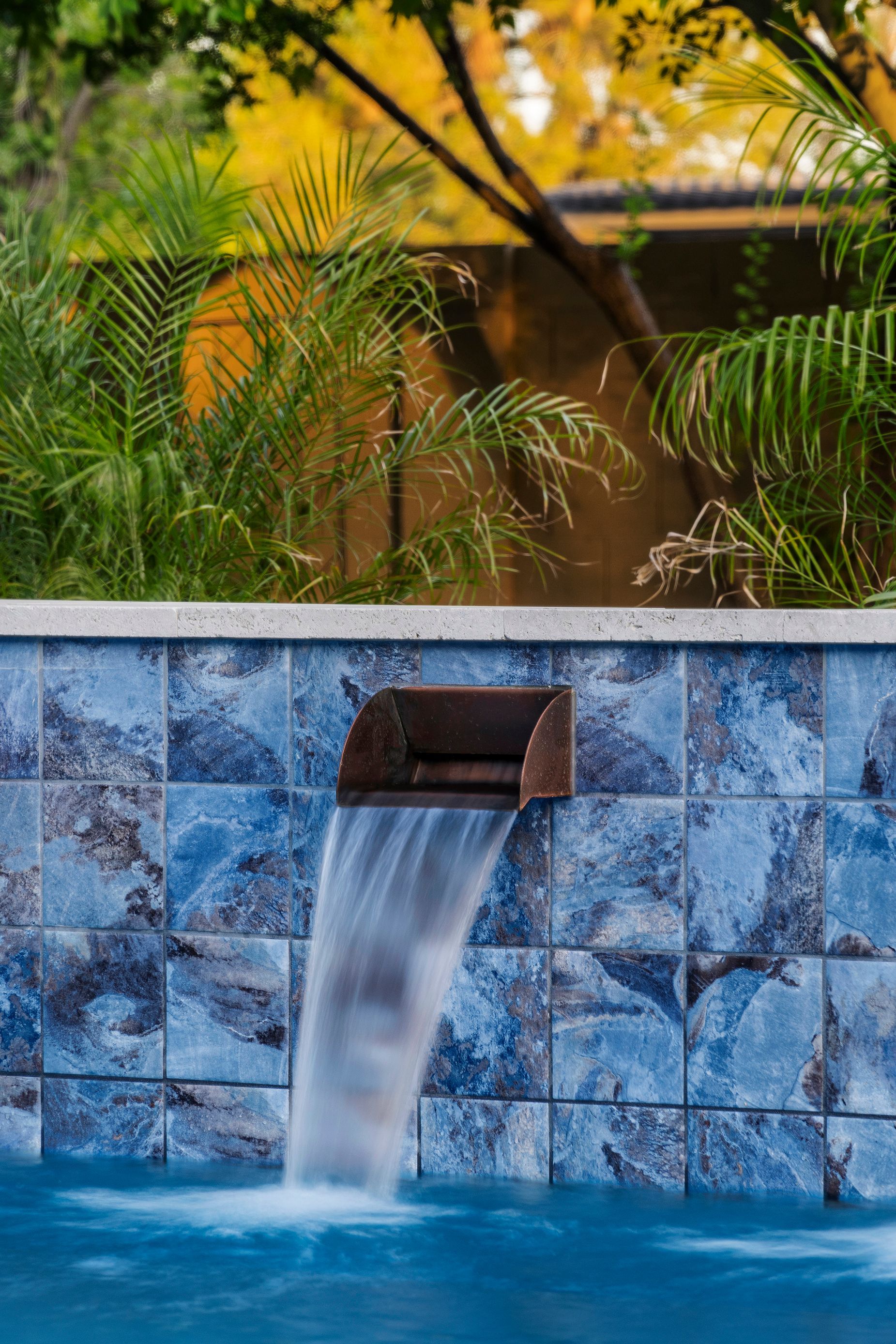 Waterfall feature over a blue-tiled wall into a swimming pool, with tropical foliage in background.