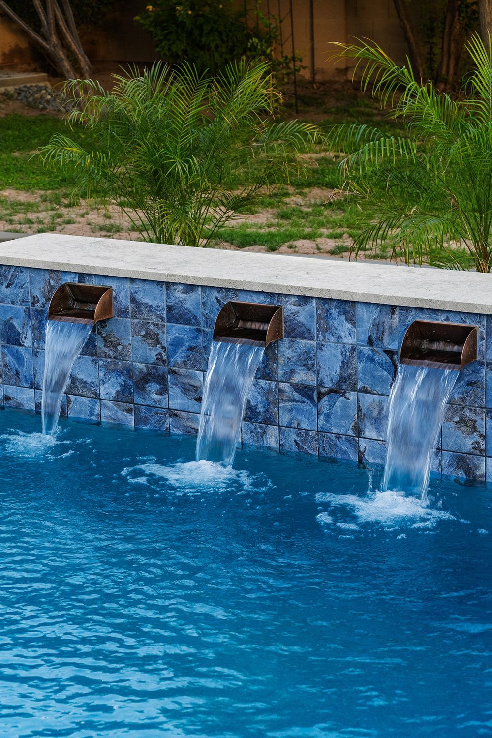 Three water fountains cascading into a blue swimming pool, against a blue tile wall, with greenery.