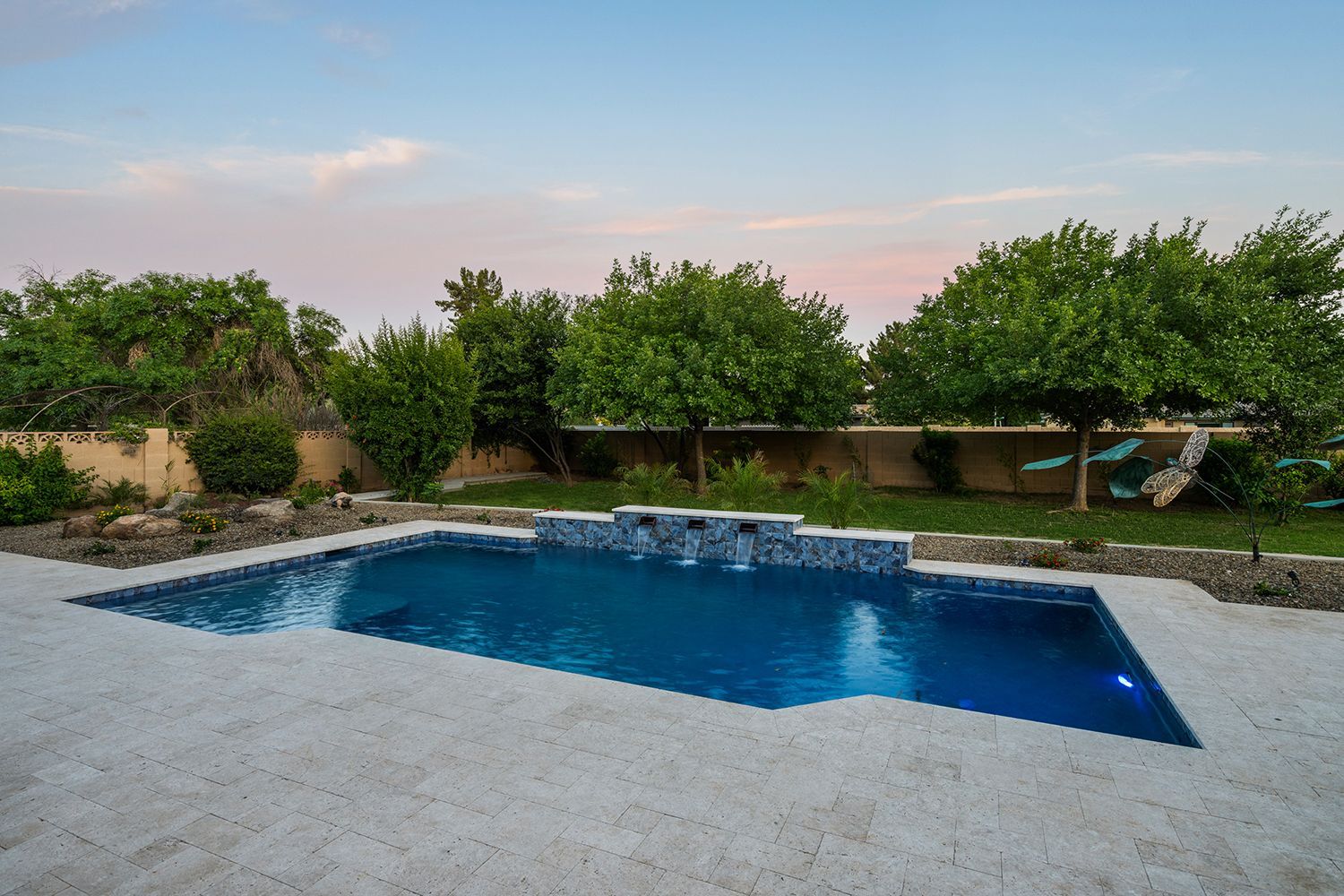 Backyard pool with stone patio, small waterfall, and trees under a colorful sky.