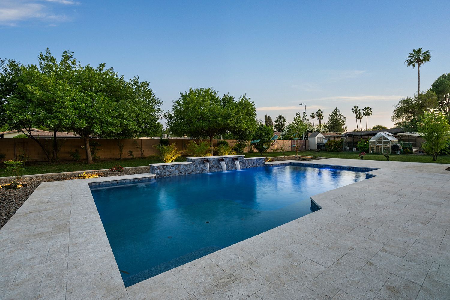 Backyard pool with blue water and stone surround; trees and greenery under a blue sky.