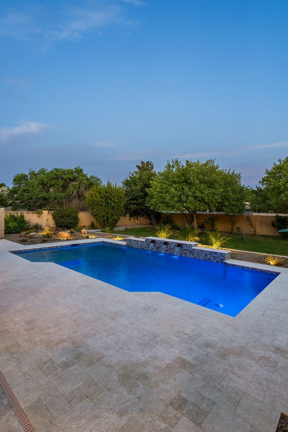 Blue-lit pool at dusk surrounded by stone patio, lush greenery, and a clear blue sky.