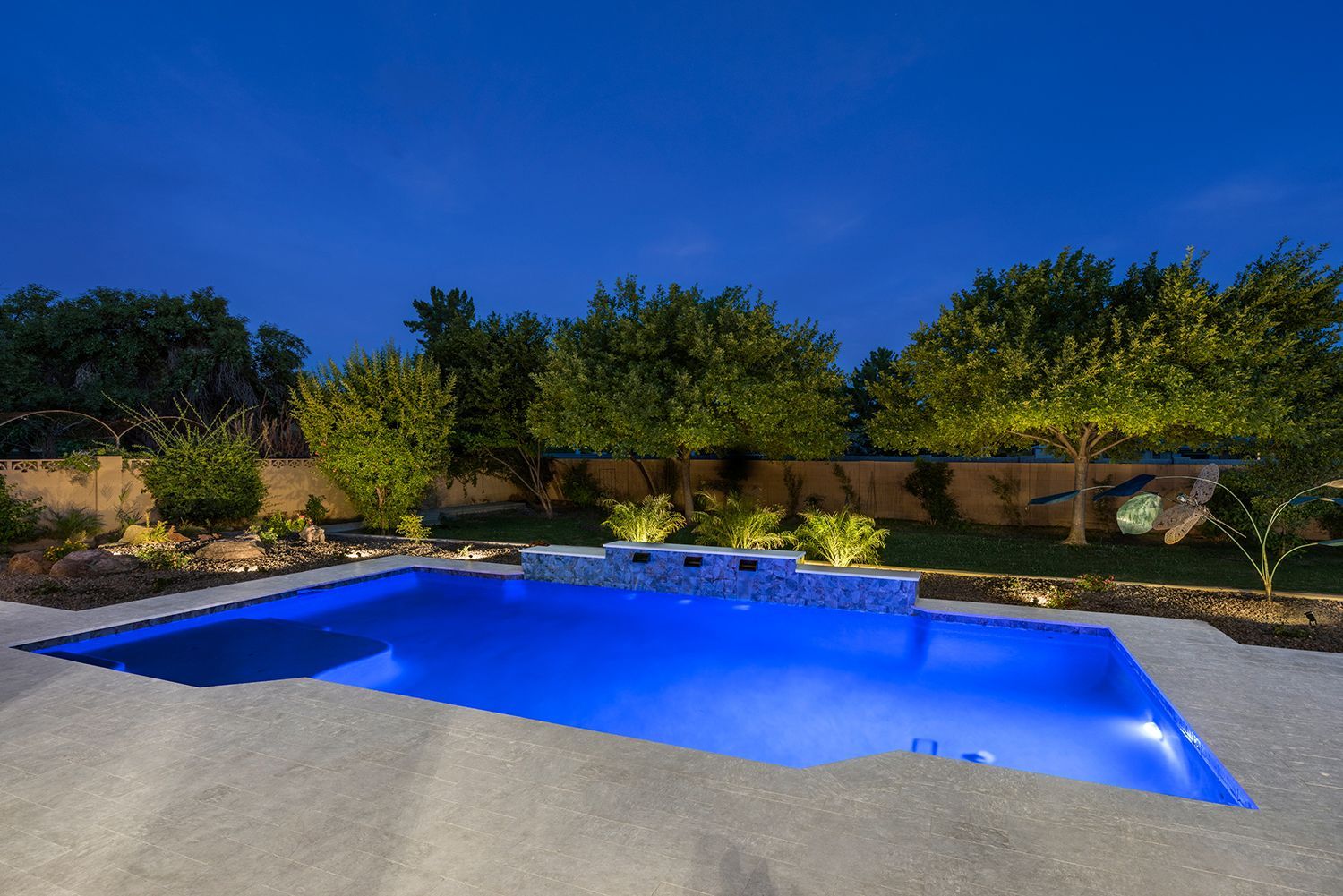 Pool illuminated at night in a backyard with trees and a stone wall.