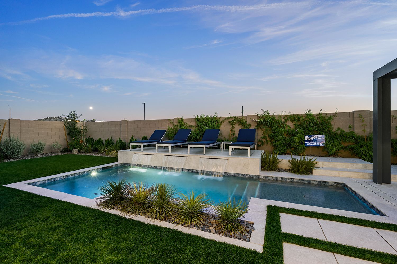 Backyard pool with lounge chairs, blue water, green grass, and a beige wall under a blue sky.
