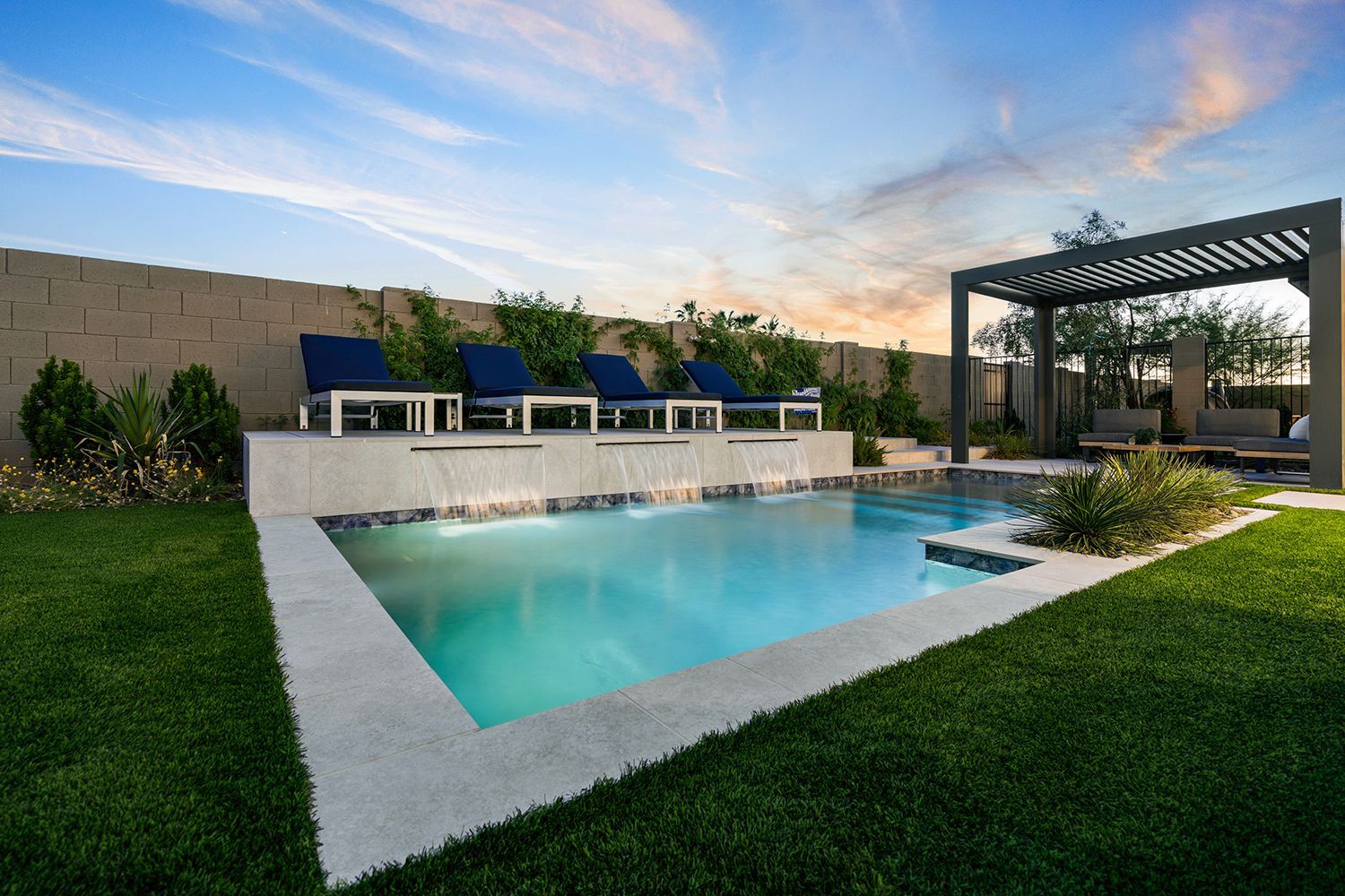 Backyard pool with waterfall, lawn, seating, pergola against a sunset sky.