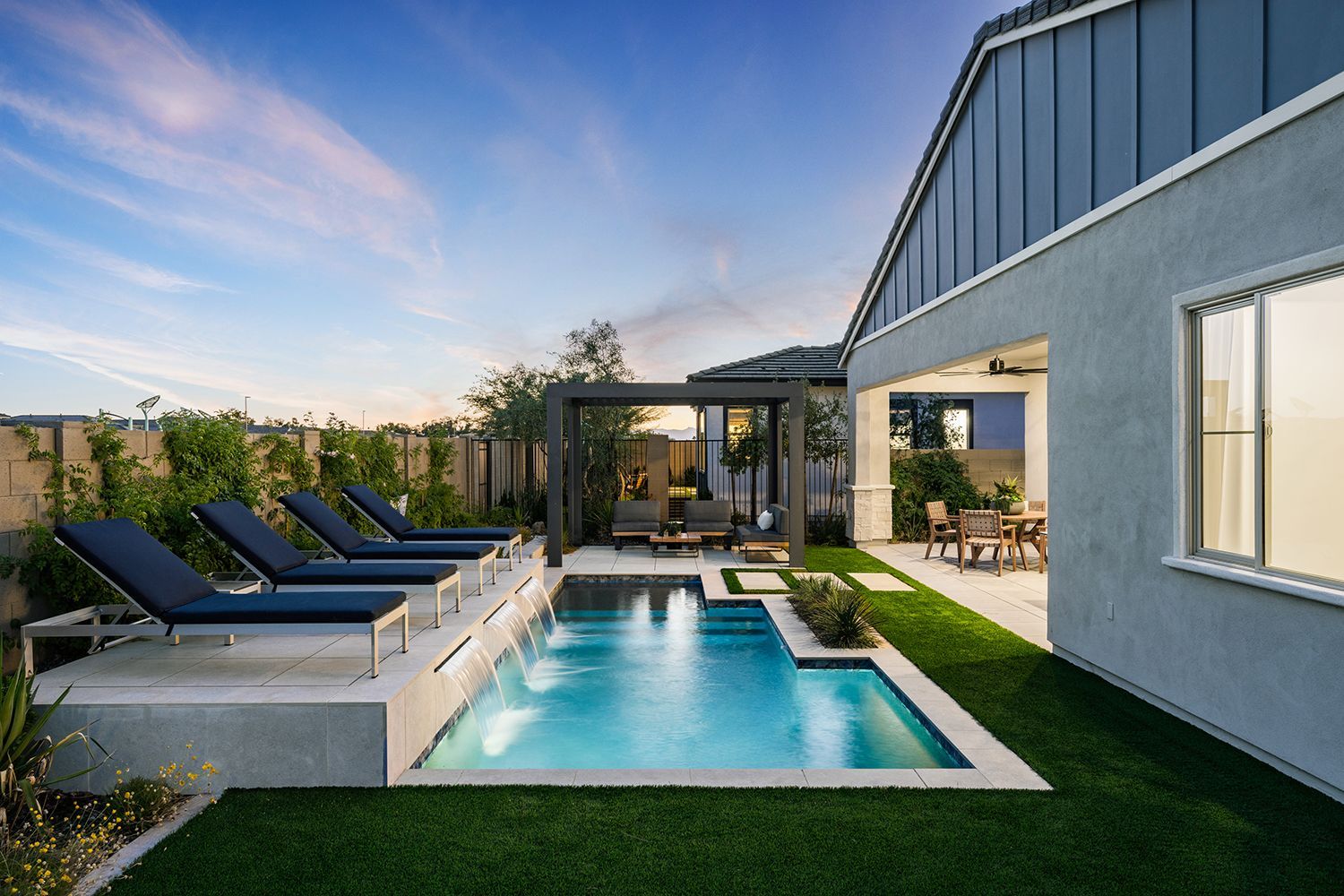 Backyard with a pool, lounge chairs, patio furniture, and a house. Blue sky at sunset.