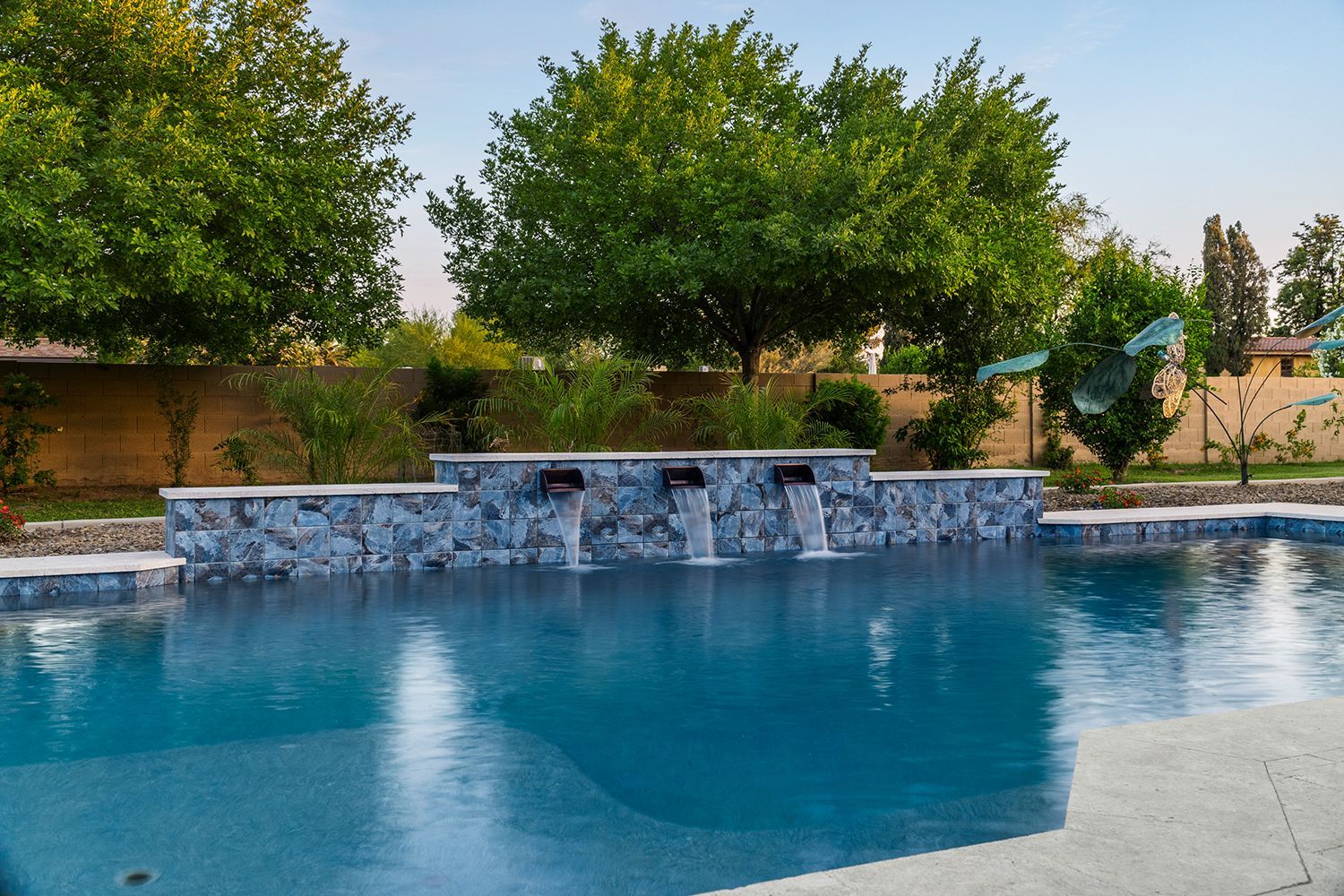 Swimming pool with water fountains against a backdrop of trees and a beige wall.