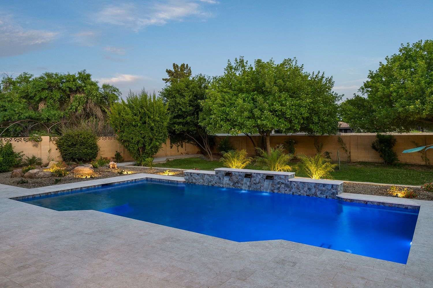 Swimming pool in a backyard, with trees and a blue-tiled wall. Pool deck, and grass surrounding.