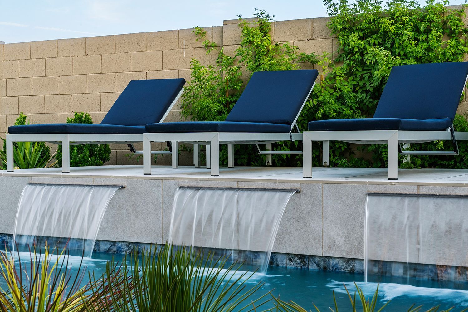 Three blue-cushioned lounge chairs on a pool deck with waterfalls; beige stone wall and lush greenery in background.