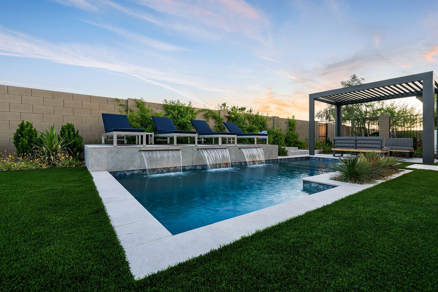 A luxurious backyard pool with water feature, lounge chairs, and pergola under a blue sky.