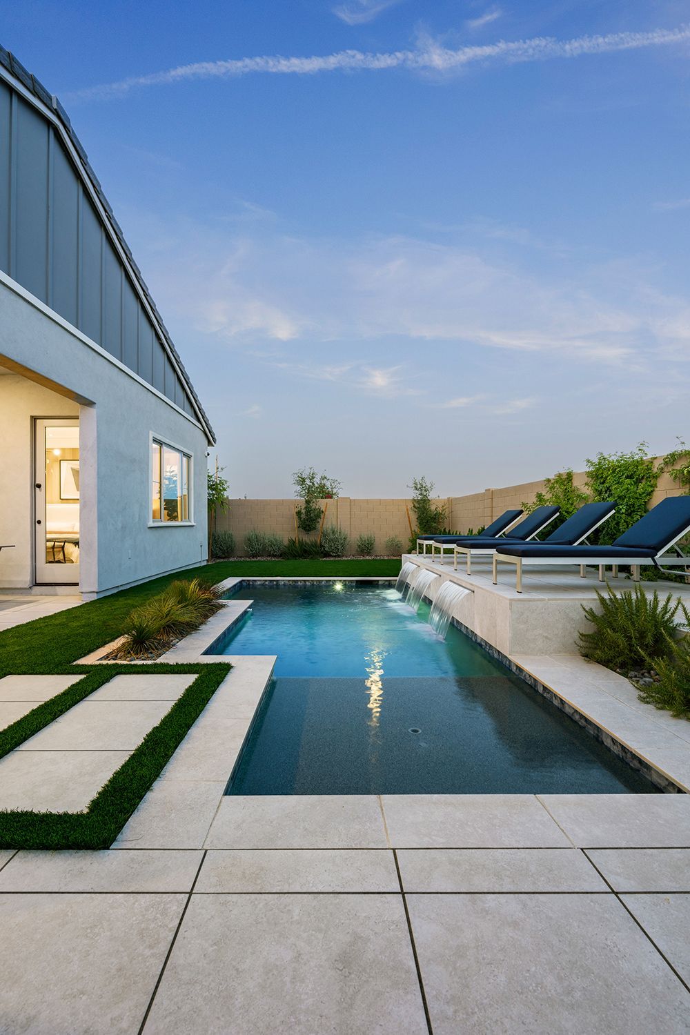 Backyard pool with lounge chairs, blue water, and modern light-colored patio; house in background.