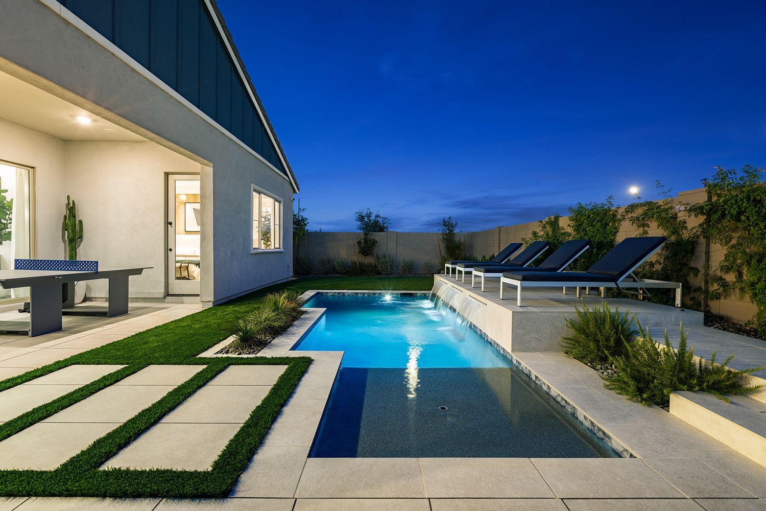Backyard with a pool at dusk; loungers, grass, patio, and modern house.
