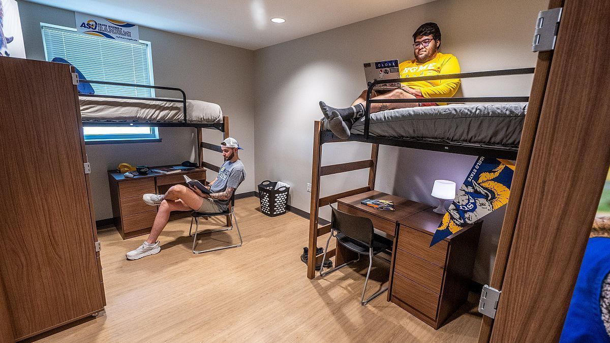 Two students in a dorm room, one on a bunk bed, the other sitting below. The room has bunk beds, desks, and closets.