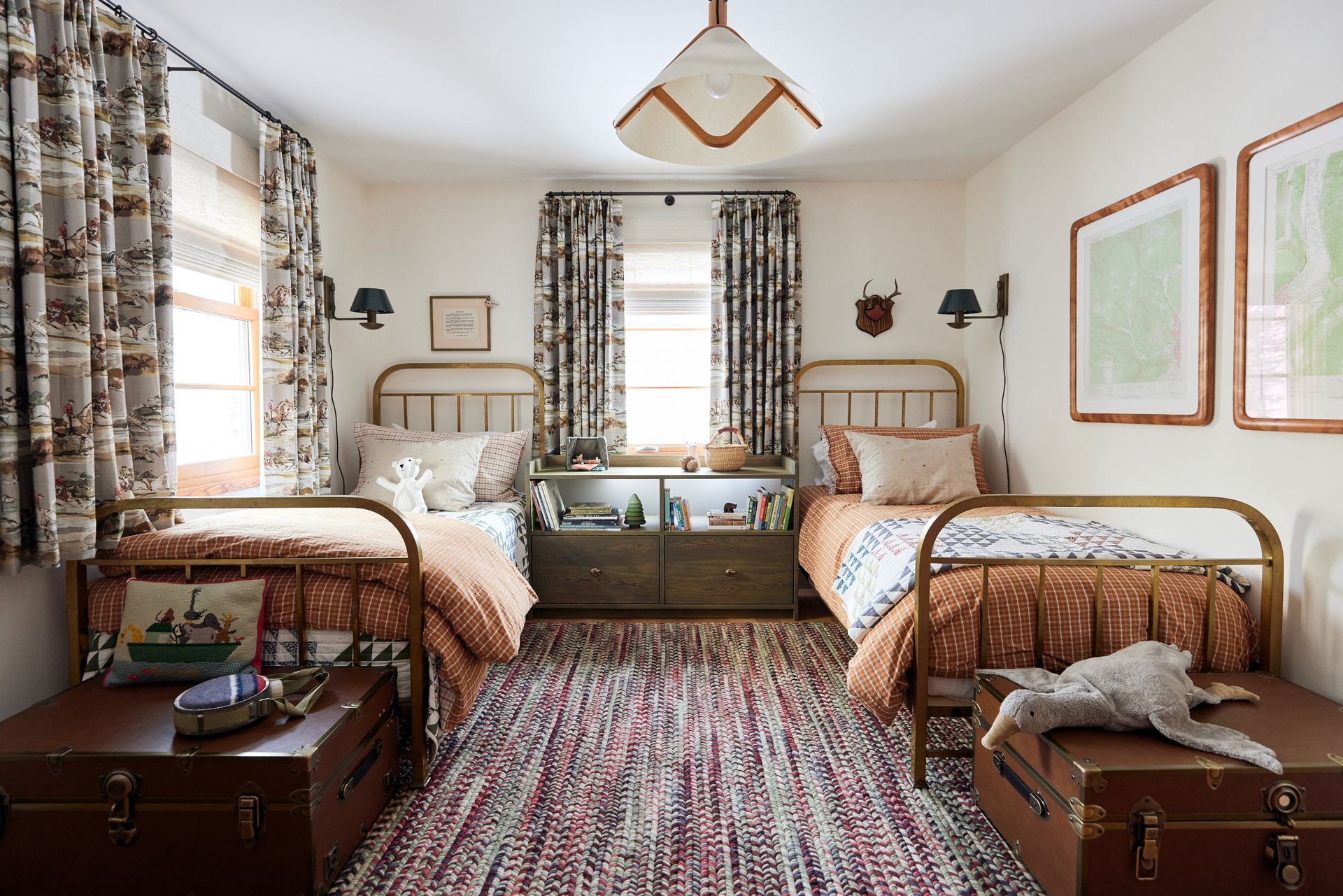 Two twin beds in a kids' room with matching brown trunks at the foot, colorful rug, and patterned curtains.