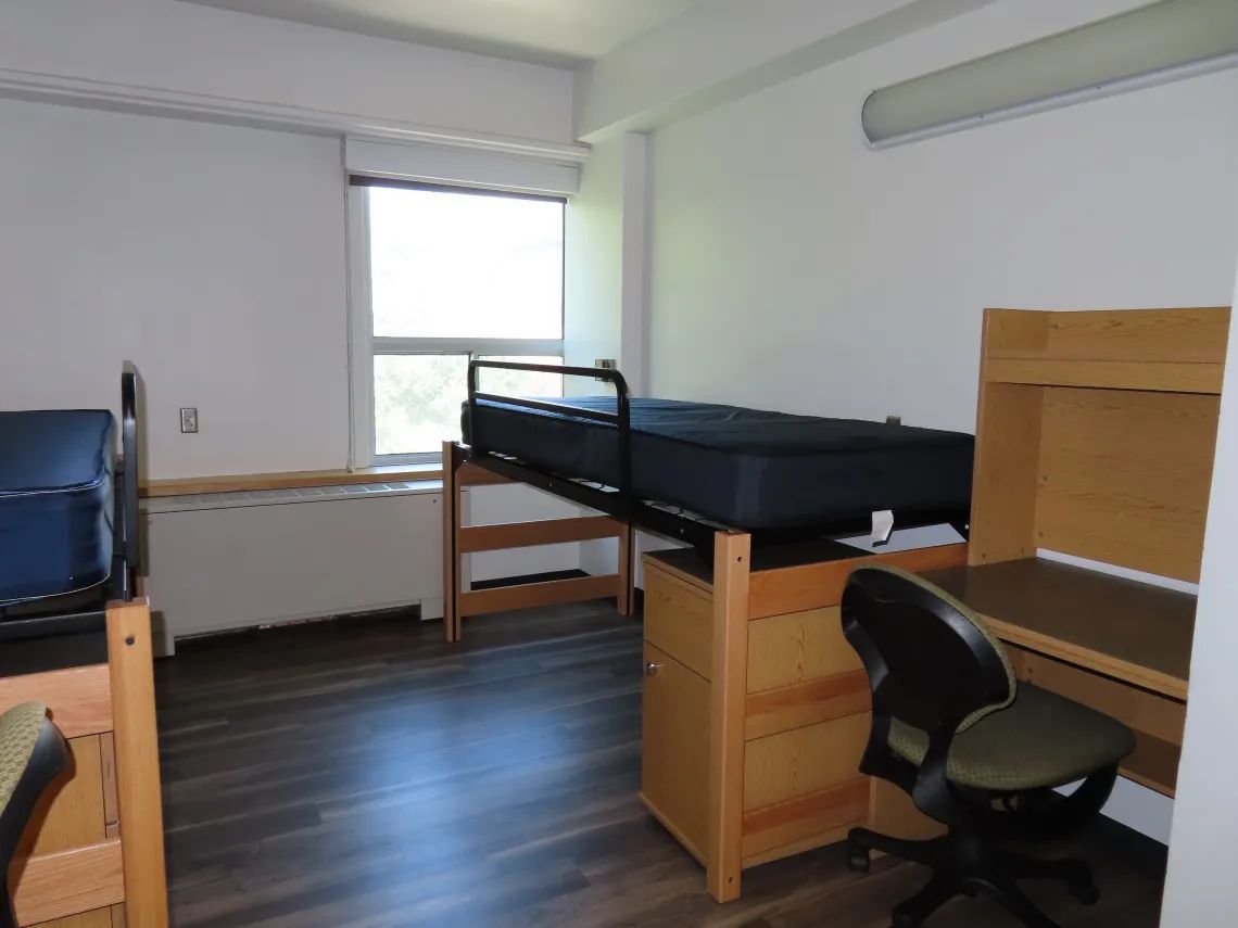 A dorm room with a bed, desk, chair, and window. Wooden furniture against white walls. Dark wood-look flooring.