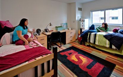 Two college students in a dorm room, one seated on a bed, the other lounging on hers. The room has two beds, desks, and an ASU rug.