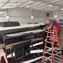 A man is standing on a ladder working on a rv in a warehouse.