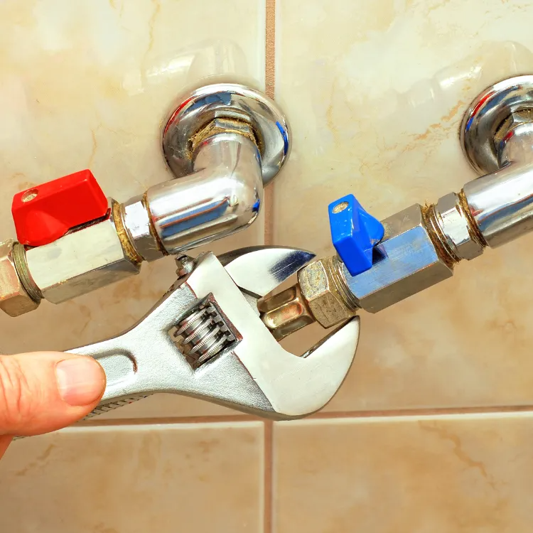 A hand uses an adjustable wrench to tighten a nut on a pipe connection next to blue and red shut-off valves.