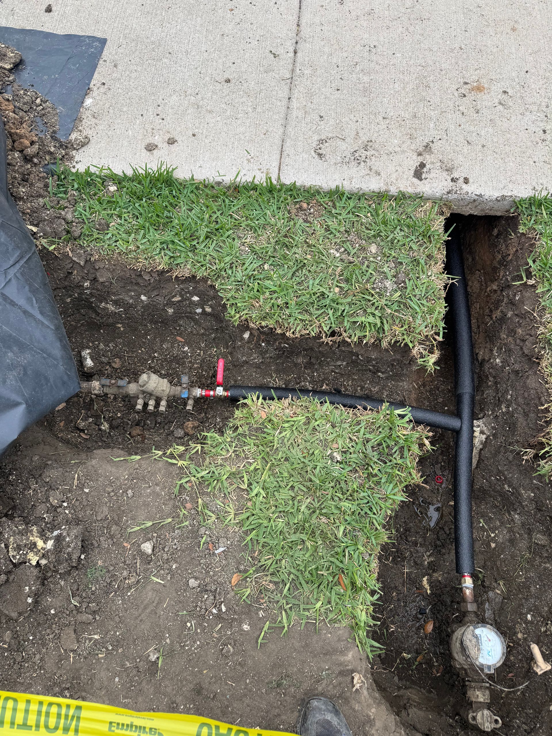 Broken sidewalk with exposed utility trench and pipe beside a grassy patch; caution tape visible at bottom left
