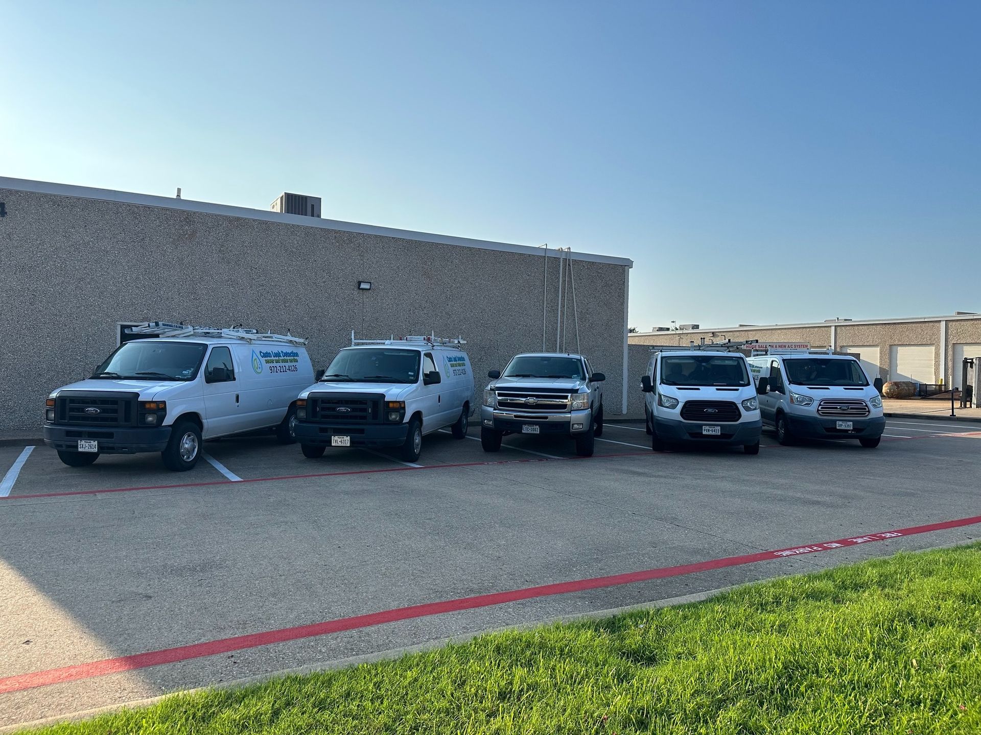 Four white work vans parked in a row against a beige brick building under a clear blue sky.