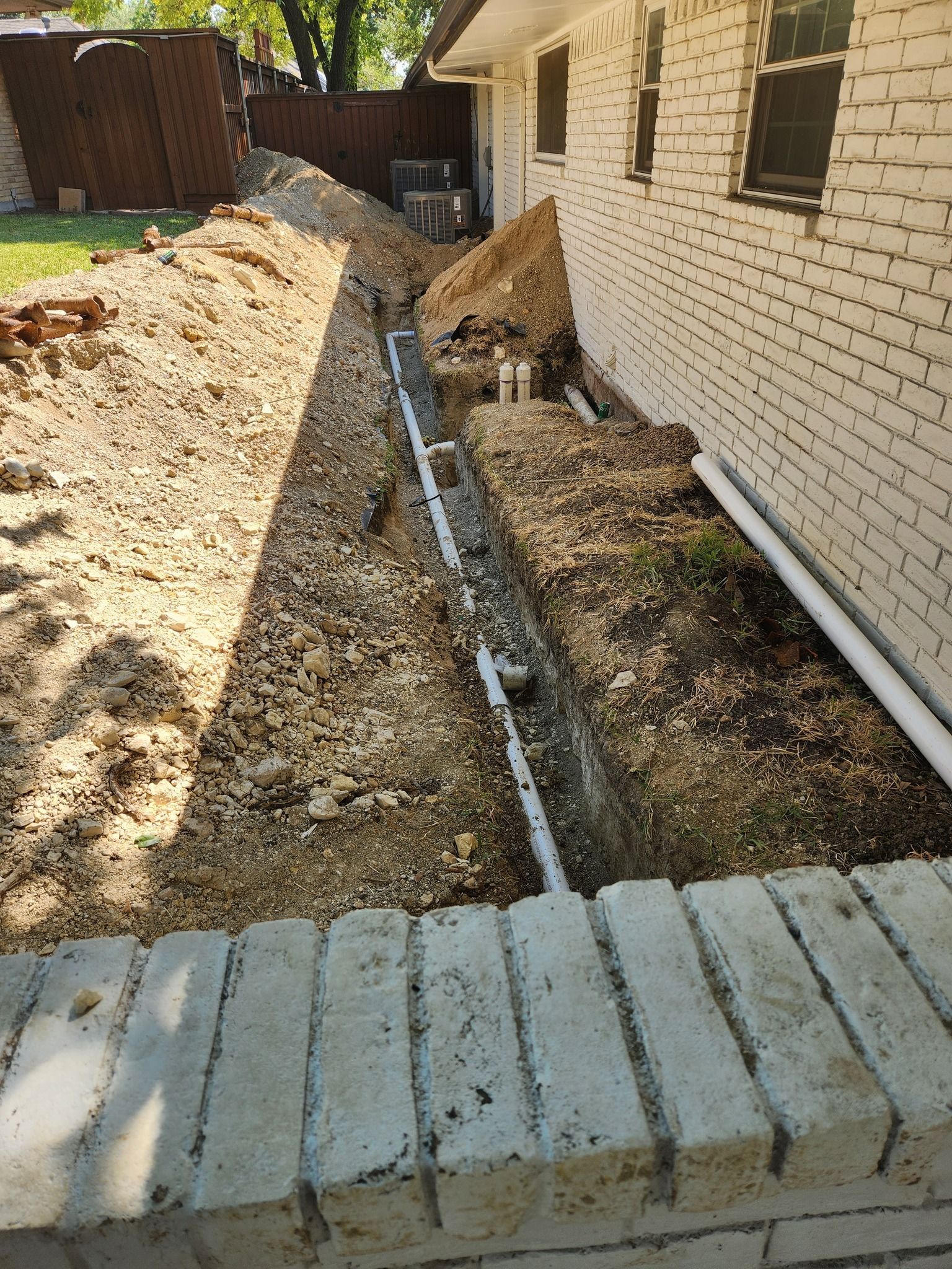 A construction trench containing a white PVC pipe runs along the side of a beige brick house.