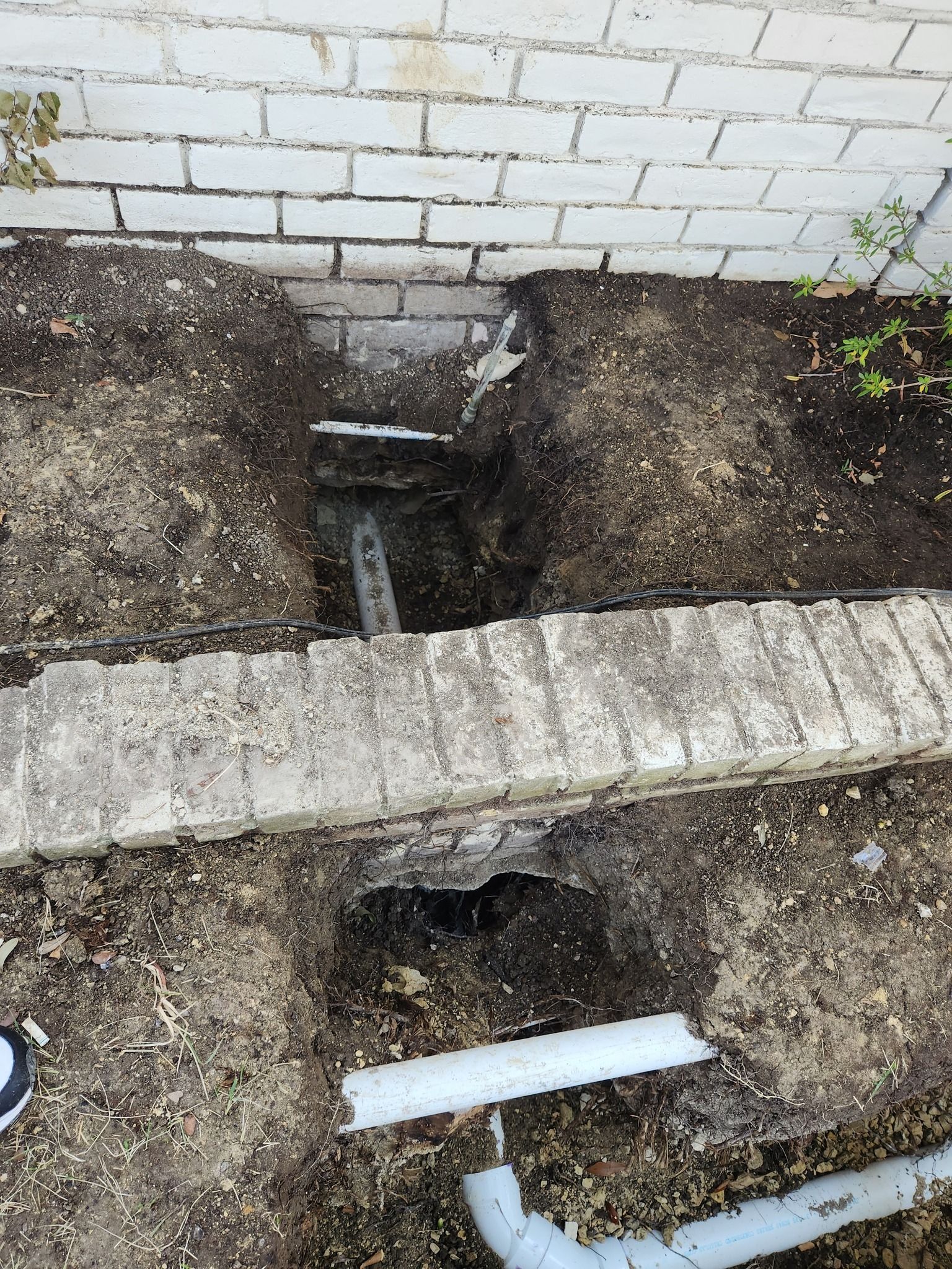 Two trenches dug in garden soil, revealing white PVC drainage pipes against a white brick wall and a low brick ledge.