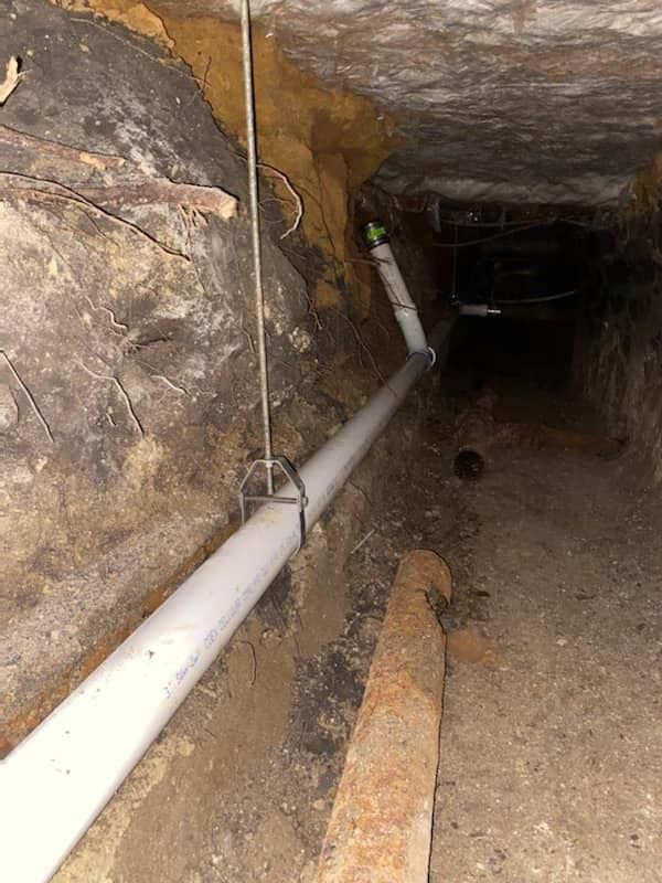 A white PVC pipe runs along a dirt wall in a crawl space, suspended by a metal hanger attached to a vertical rod.