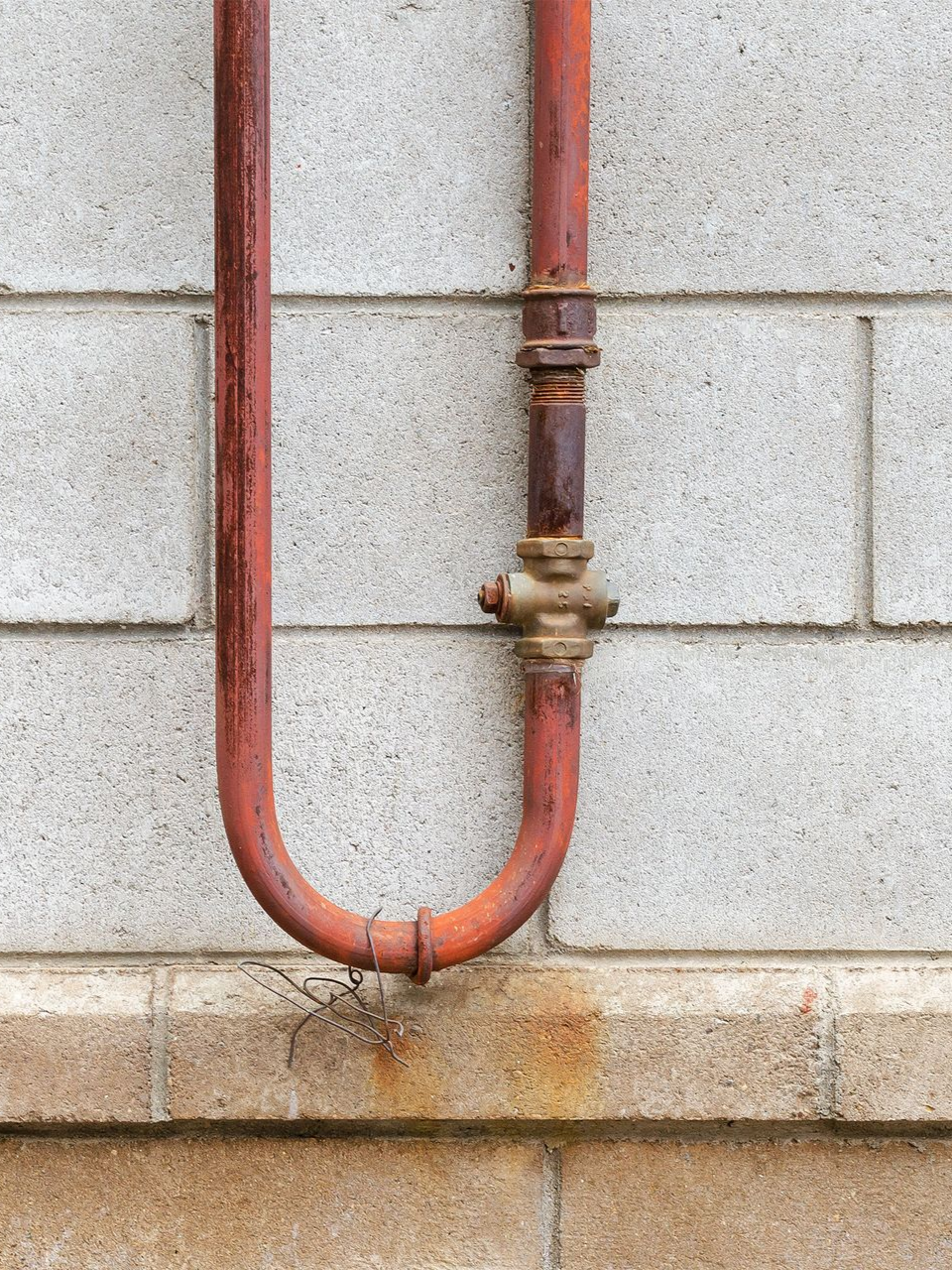 A U-shaped rusty red metal pipe with a brass-colored valve, mounted against a textured gray cinder block wall.