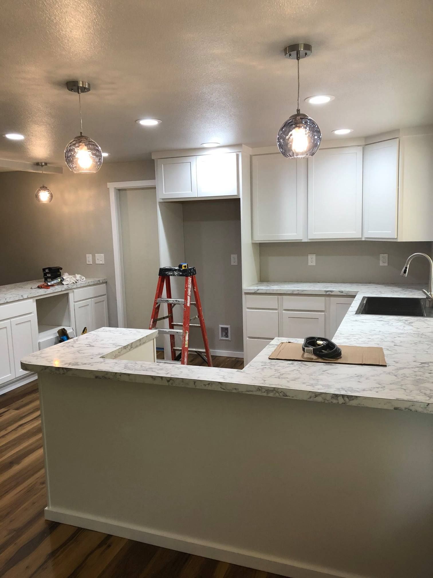 Newly renovated white kitchen with granite countertops, recessed and pendant lighting, and a ladder.
