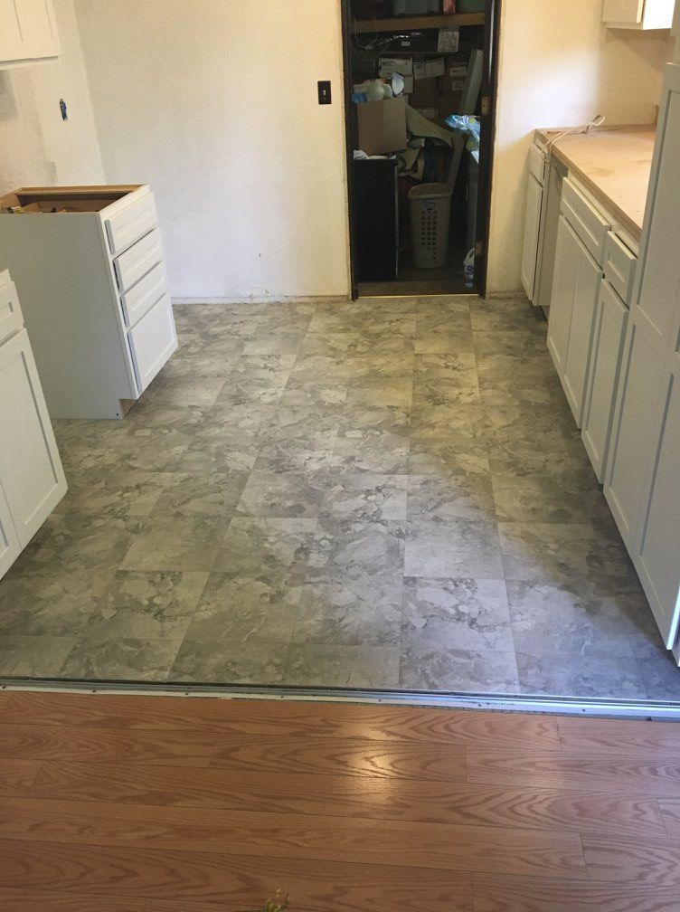 Kitchen remodel in progress: white cabinets, gray tile floor, doorway to storage area, brown wood floor at edge.