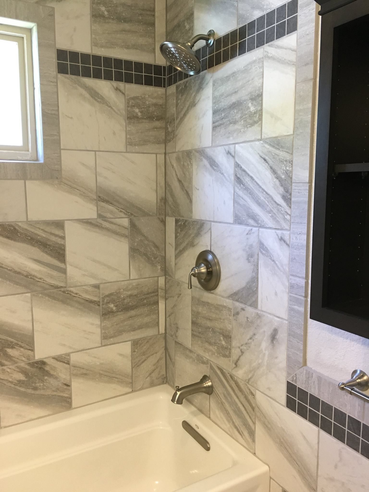 Bathroom with white bathtub and shower tiles. Gray and white veined tiles and black decorative strip.