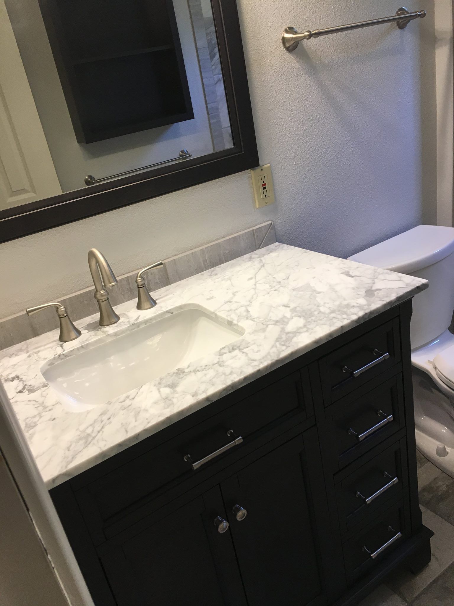 Dark vanity with white marble countertop, sink, and faucet in a bathroom. Toilet is visible.